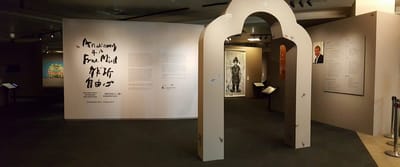 A photo of the exhibition entrance. In the foreground, there is a narrow archway shaped like a bell. In the background, there is the informational wall.