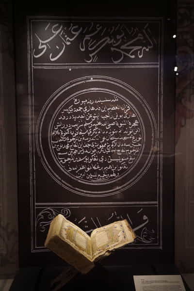 A tall showcase with the Qur'an, opened up to illuminated manuscripts. A black and white graphic with similar illuminated manscript decorates the back of the showcase.