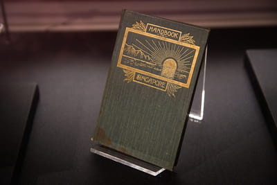A dark green book is propped up, and its title and illustration is gilded in gold leaf. The title is 'Handbook to Singapore'. The illustration is of a sun halfway in the sea, with mountains on the side.