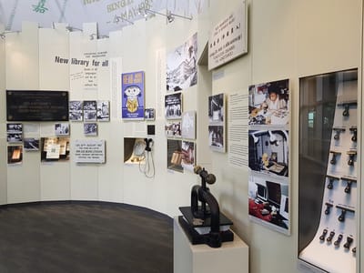 Various photographs are on display, along with glass showcases of book stamps and due date cards. An old book-press machine is on a pedestal. Next to it on a wall, there is a small TV screen with audio handsets.