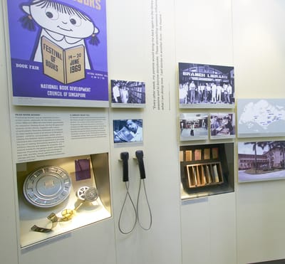 A photo close-up of a wall display, featuring the 1960s history of the National Library. On the top left, there is a poster with a cartoon girl and her book, by the National Book Development Council of Singapore. In the center, there is a small TV screen with audio handsets. On the right showcase, there is a wooden box with old due date cards on display.