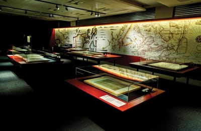 Several table showcases featuring maps are in a room, with a giant map graphic on the back wall.