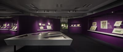 A section of the exhibition, with purple wall showcases and a silver table-top showcase in the middle. Maps are on display.