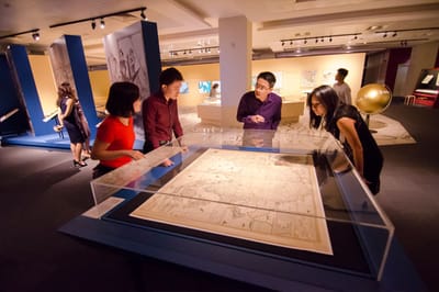 A table showcase with a map is surrounded by a curator and his tour group. Some of them are looking at the map, while the others are listening to the curator's talk.