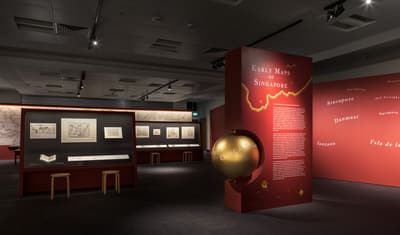 Several red walls line this exhibition section. In the middle is a title wall labelled 'Early Maps of Singapore', with a gold globe half-embedded on its side. Maps are displayed up on the walls.
