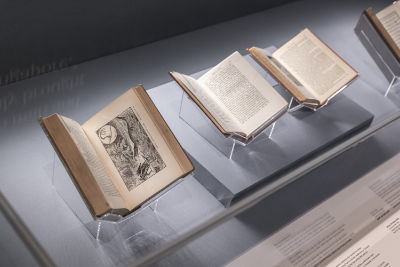 Several books are on display in a showcase. A book is opened up to a black and white etching illustration of several men pulling a rope around a crocodile.
