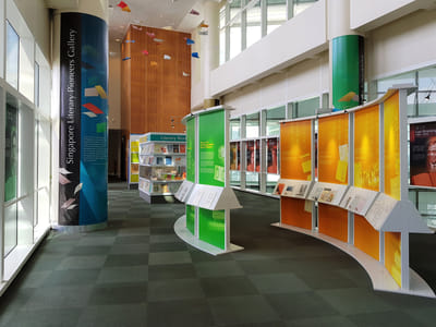 Green and orange curved walls with information panels on them are in the middle of the exhibition. Tall banners cover the pillars at the sides. A bookshelf is in the back.