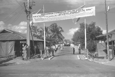 A black and white photo of a big banner hung across a newly paved road between two posts. It announces the official opening of Jalan Nanas (Nanas road).
