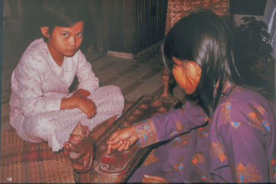 Two Malay girls dressed in their baju kurung (a traditional Malay costume worn by women) play a game of congkak (a traditional Malay two player game in which seeds or marbles are dropped into carved holes in a wooden board) on the floor.