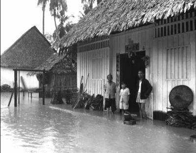 Three individuals stand in floodwater at the doorway of their kampong home.