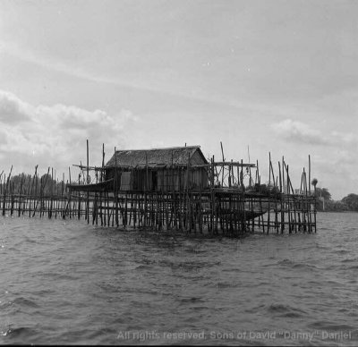 A black and white photo of a kelong (offshore fishing platform), consisting of a thatched house built on stilts. It stands in the midst of calm sea waters.