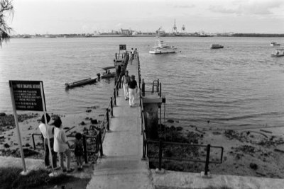 A black and white photo where a handful of visitors explore the Punggol jetty while some boats lie on the water a short distance away to the right.