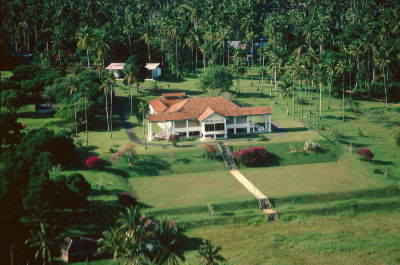 An aerial shot of Matilda House, a single-storey colonial bungalow with a red roof and a black-and-white façade. The bungalow stands amidst spacious green lawns, surrounded by a forest of coconut trees.
