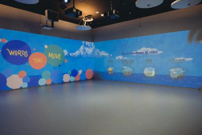 A photo of the Words That Move room. It's empty, and the left wall has the Words That Move title projected onto it. On the right wall, four poems are available for selection.