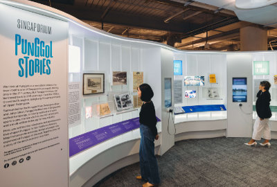A photo of the Singaporium space. An introduction panel is on the left, and showcases and multimedia screens are on its right. A visitor is looking at the displayed items in a showcase, and another visitor is browsing the multimedia screen while holding its sound handset.