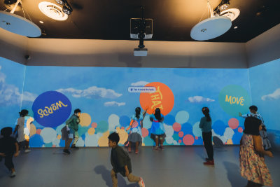 A photo of the Words That Move exhibition. Graphics of colourful bubbles, with the title 'Words That Move' on three individual bubbles, are projected onto the walls. Children are either touching the walls or running around.