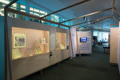 A photo of an exhibition section. On the left wall, wall showcases with books and items are inside. There is a small TV screen displaying an image.