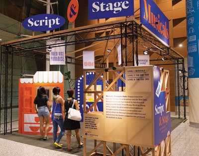 A photo of the lobby exhibition: Script and Stage - Musicals. Three people are entering the exhibition. It is shaped like an open box, supported by black metal railings. Small banners hang from its ceiling, and small wall displays are inside the exhibition.