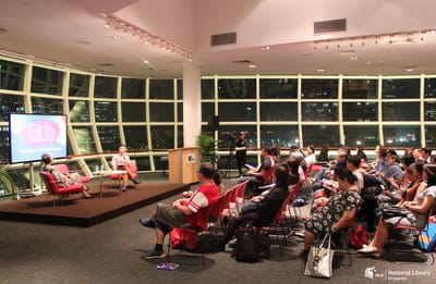 A photo of the event, with an audience seated on the right. On the left are the woman and man on stage.