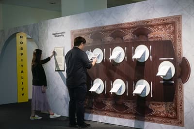A photo of the Gastronomic Diversity section, with a woman and man browsing it. The wall has a giant dinner table with dinner plates. Informational 'menus' are mounted onto these plates.