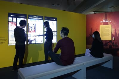 There are two people browsing 3 tall multimedia screens on the wall, with two more people sitting on the bench behind them. On their right, a wall is titled 'Epic Journeys'.