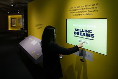 A woman interacts with a screen embedded into a wall, with a RFID reader slot below it. The wall reads: 'Scan the tags you picked up at each department in the scanner below to vote for your favourite parts of the exhibition'.