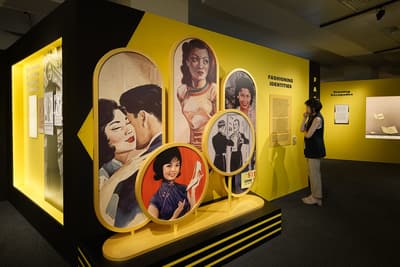 A photo of the Fashioning Identities section, with a woman looking at the information text. In the foreground, there are several circular stands featuring illustrations of glamourous women and men.