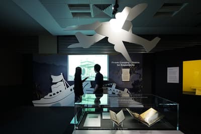 A photo of the From Luxury Liners to Superjets section, with a man and woman looking at a projected image in the background. Cut-outs of paper plans are hanging from the ceiling. In the foreground, there is a showcase featuring images and books.
