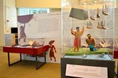 There is a small showcase with paper sculptures, sitting on top of an audio kiosk. It is of a man by the riverside holding a boulder and several ships aloft in the air, while a Sultan bows before him.