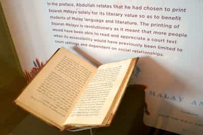An opened book sits within a tall showcase. It contains Jawi script. In the showcase background, there is a printed graphic with a write-up about the Sejarah Melayu on it.