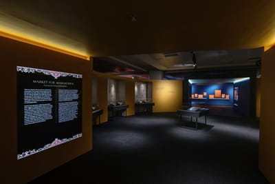 A corridor leads into a room. On the corridor's left wall, a lightbox display is titled: 'Market for Manuscripts'. In the room inside, a showcase is in the center, surrounded by other showcases against the walls.