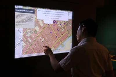 A man touches an interactive screen on the wall. The screen displays a city map with several map markers, along with the information box titled: 'Early Malay Muslim Printers'.